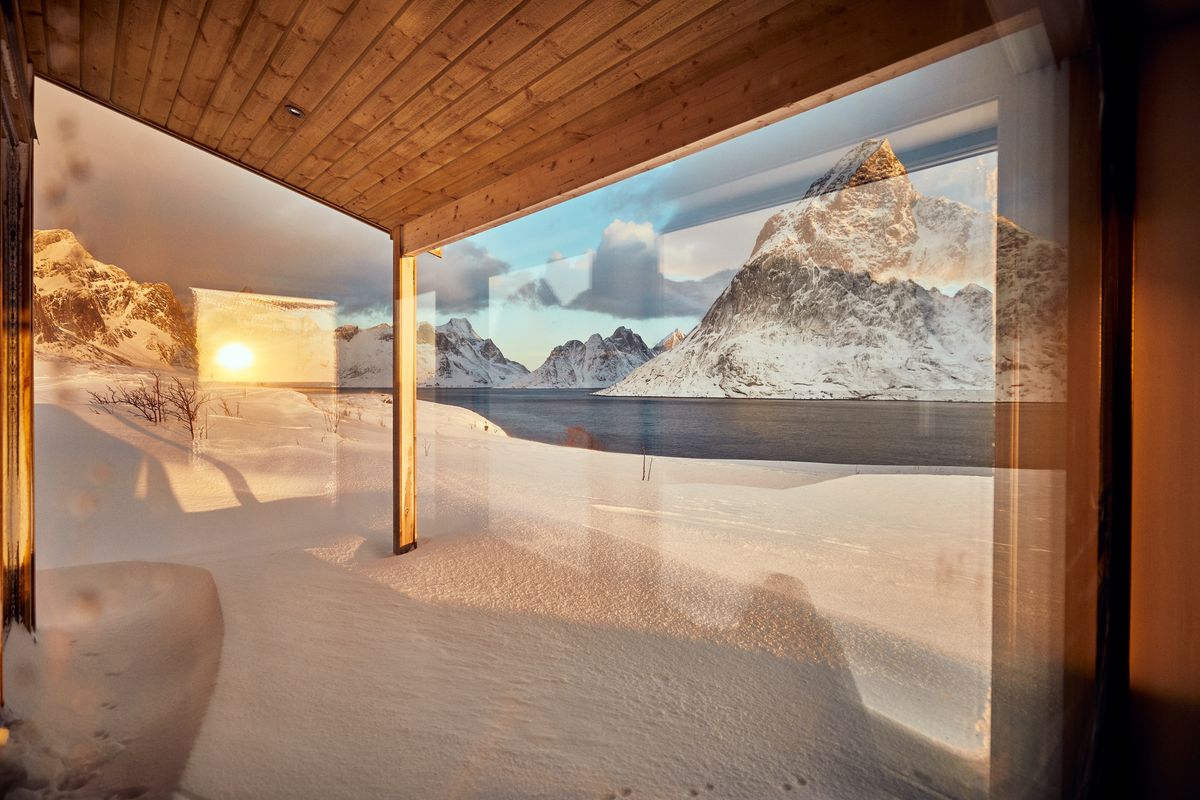 View from under the wooden roof of the Reine house through glass onto deep snow, the fjord and a sharp peak in low sun