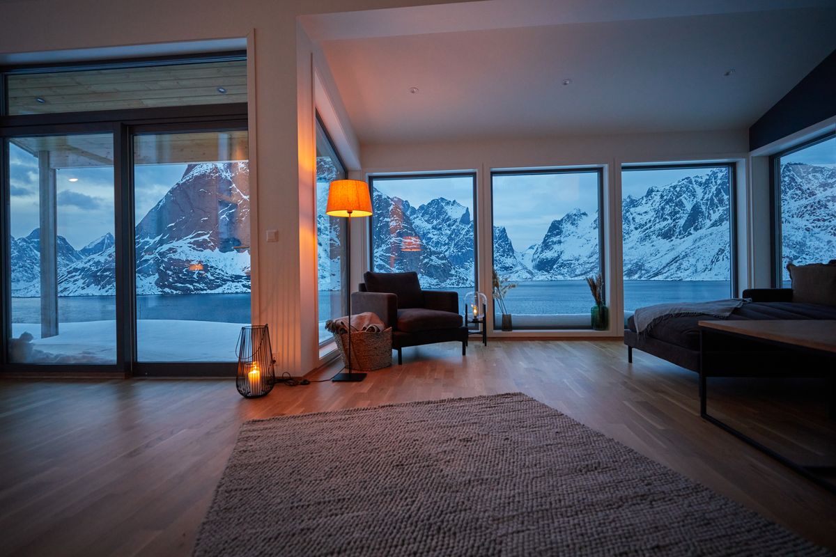 Living room in Reine at dusk, warm lamps inside and snowy mountains behind the glass