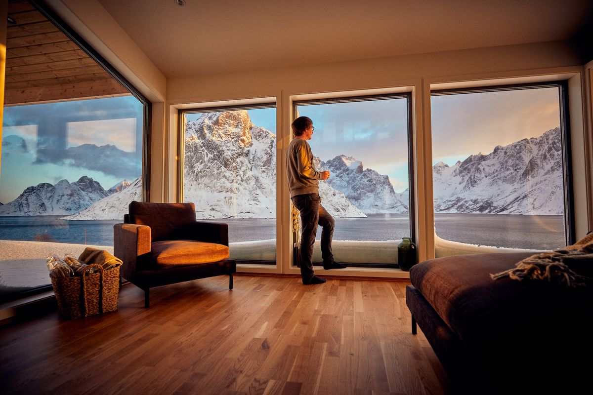 A man stands at the tall windows of the Reine living room, snowy peaks across the fjord