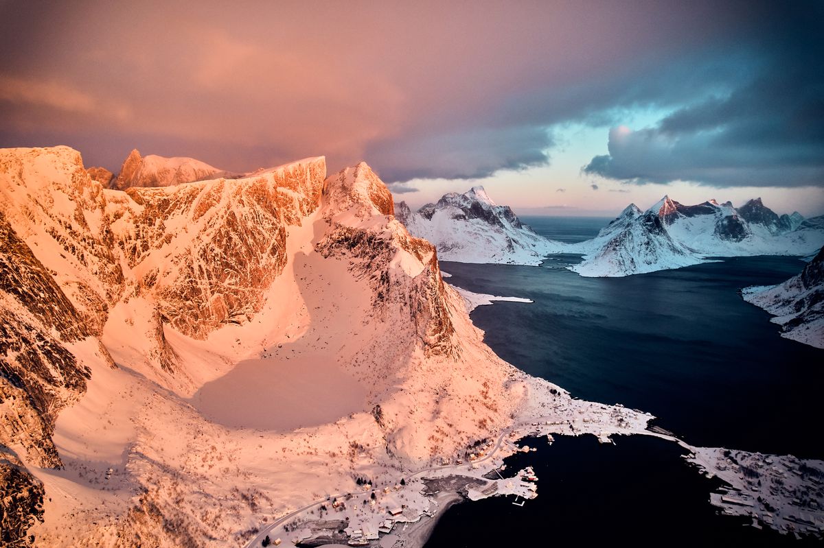 Snow-covered peaks above Reine glowing pink in the first morning light, seen from a drone