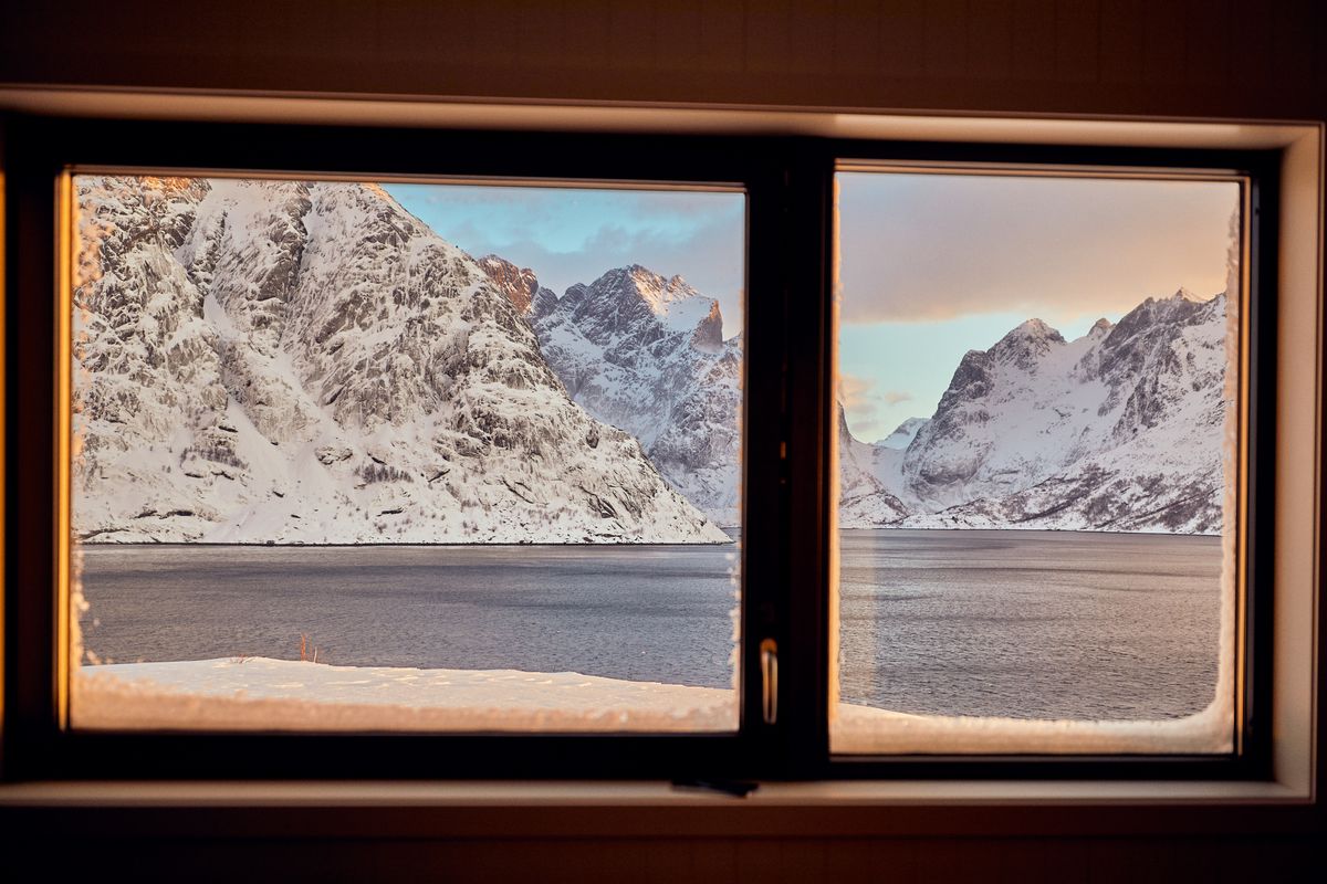 Looking through a dark window frame at a snowy mountain across the fjord